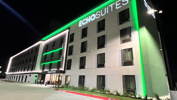A hotel named ECHOSUITES with a modern facade, illuminated green accents, and a fire lane in front.