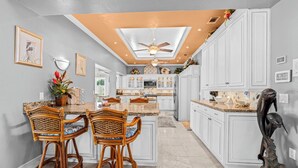 A kitchen with white cabinets, a central island with two wicker chairs, a ceiling fan, and a decorative sculpture. - VRCC Villa Dolphin House (Cape Coral)
