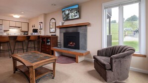 A living room with a fireplace, a television mounted above it, and a large window with a view of the outdoors. - Okemo Mountain Lodge unit with Direct Access to Clocktower Base Area at Okemo (Ludlow)