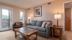 A living room with a sofa, two side tables, a coffee table, a chair, and a door. - Okemo Mountain Lodge unit with Direct Access to Clocktower Base Area at Okemo (Ludlow)
