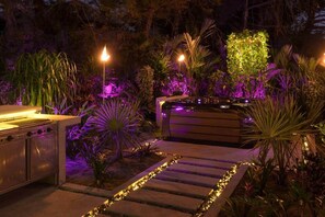 Un área de cocina al aire libre con una parrilla, rodeada de plantas tropicales e iluminada con luces ambientales.