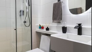 A modern bathroom with a glass shower enclosure, a white toilet, a vanity with a mirror, and a small trash bin.