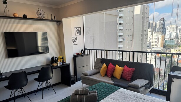 A modern bedroom with a large bed, a sofa with cushions, a desk with a TV, and a balcony with a city view.