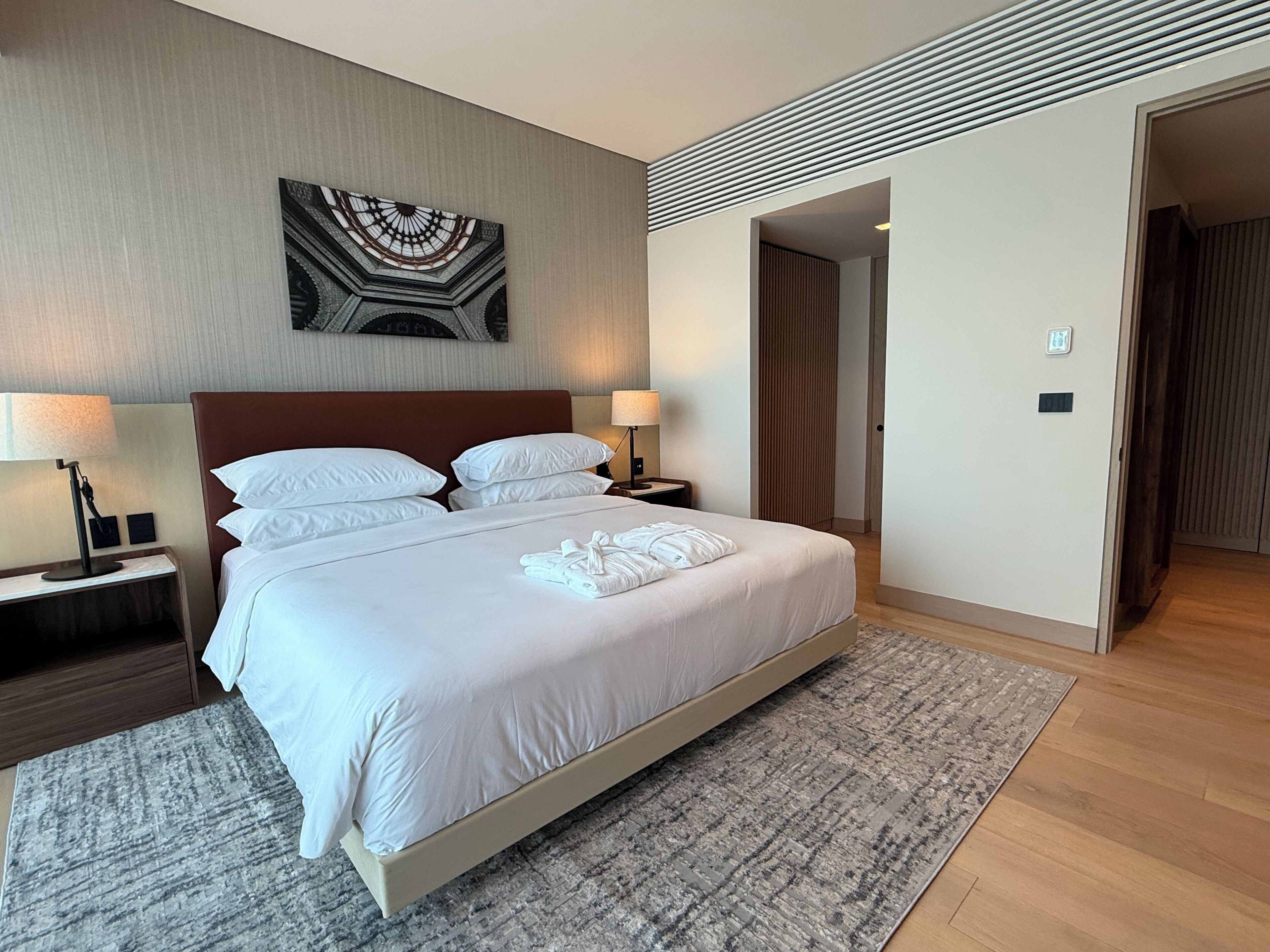 Une chambre d’hôtel avec un grand lit, deux tables de chevet avec des lampes, une œuvre d’art accrochée au mur et une porte donnant sur une autre pièce.