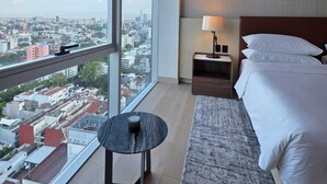 Et hotelværelse med et stort vindue, en seng, et natbord med lampe, og udsigt over byen.