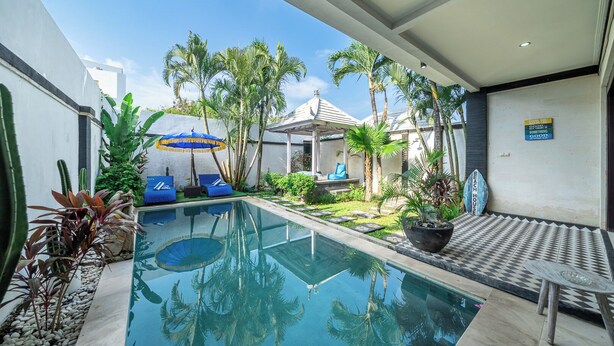 Una piscina con sillas de descanso y sombrillas, rodeada de plantas tropicales y un pequeño edificio.