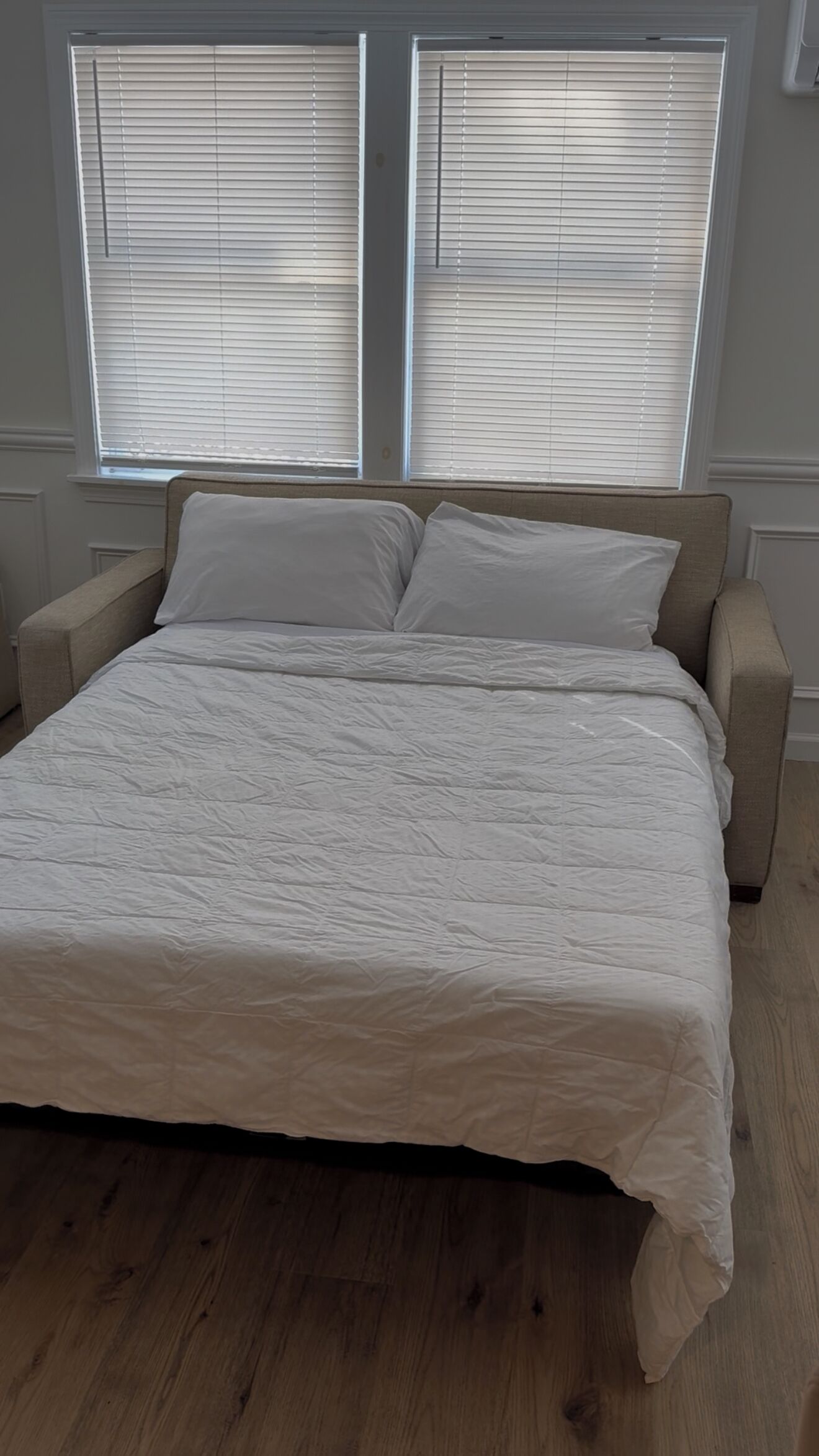 A bedroom with a bed, two windows with blinds, a wooden floor, and a white wall.