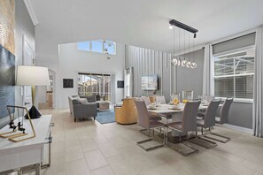 Amplia sala de estar con mesa de comedor, sillas modernas y sofá. Hay un televisor montado en la pared y una lámpara en el techo.