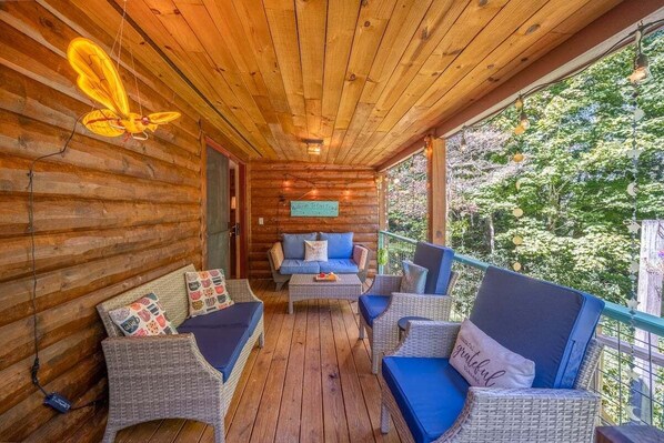 Une cabane en rondins de bois dotée d’un porche aménagé avec des meubles en osier, un canapé bleu et une pancarte de bienvenue.