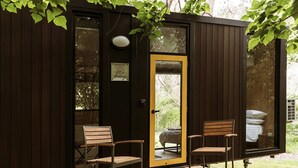 Una cabaña de madera con una puerta amarilla, dos sillas de madera y una cama visible desde el interior.