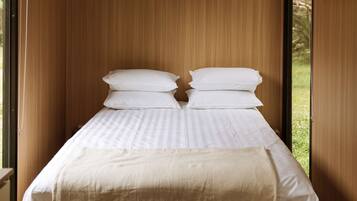 A bed with white bedding and a beige throw blanket, set against a wooden wall with a window revealing greenery outside.