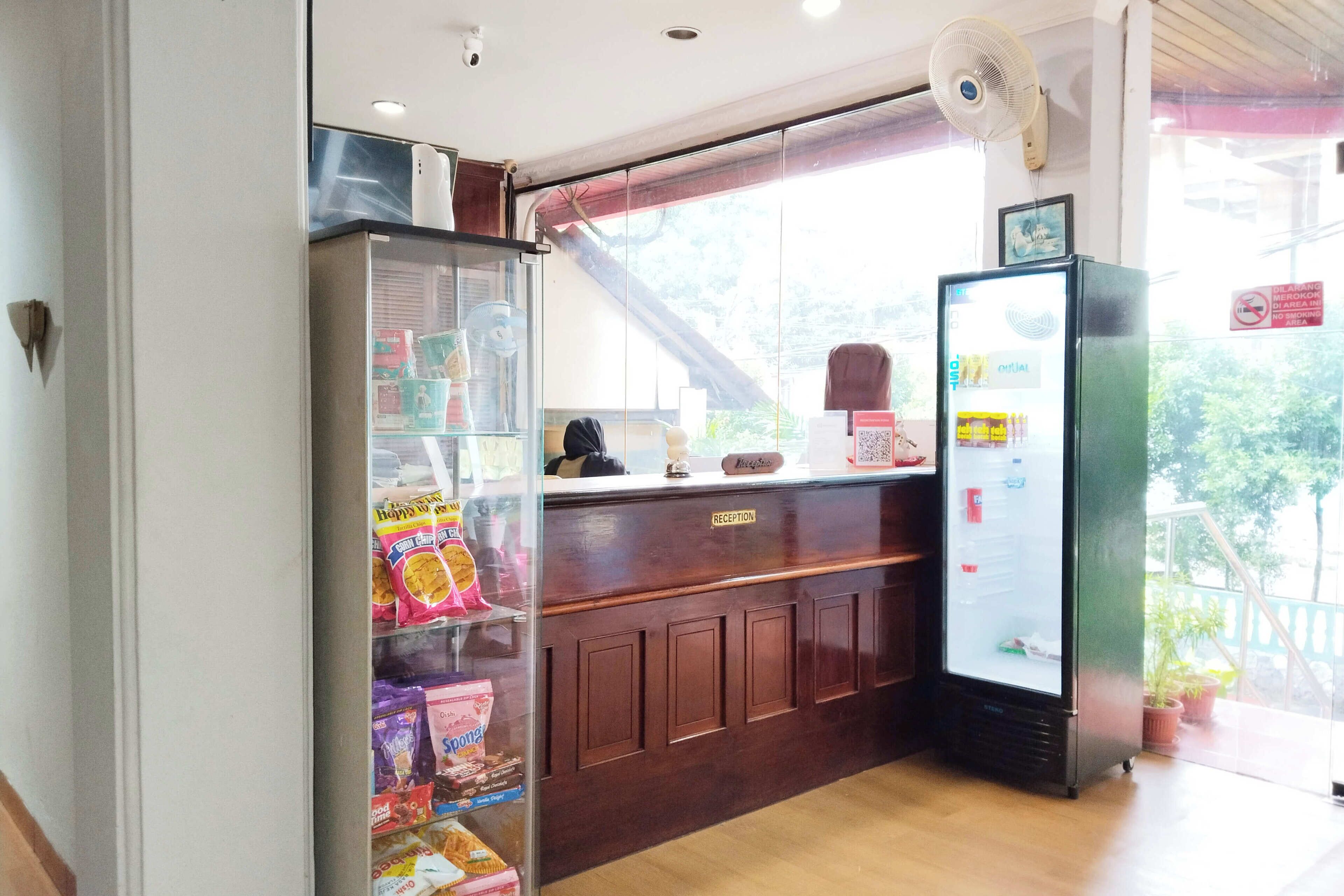 Een receptiegedeelte met een glazen displaykast vol snacks, een houten balie, een koelkast en een ventilator.