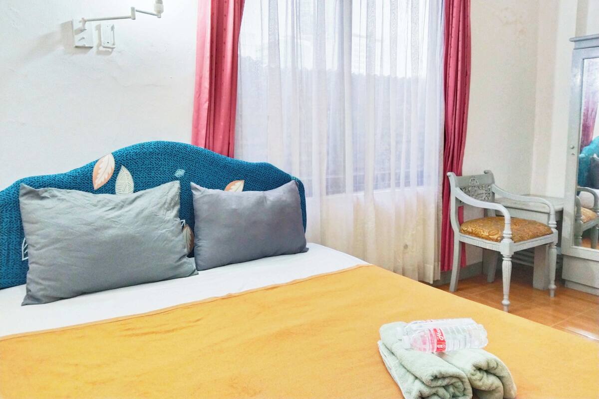Una cama con cabecera azul, almohadas grises y una colcha amarilla. Hay una silla con un cojín y una toalla doblada en el suelo.