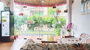 Une pièce comprenant un lit, une petite table, un ventilateur et une fenêtre donnant sur un paysage verdoyant et des bâtiments.