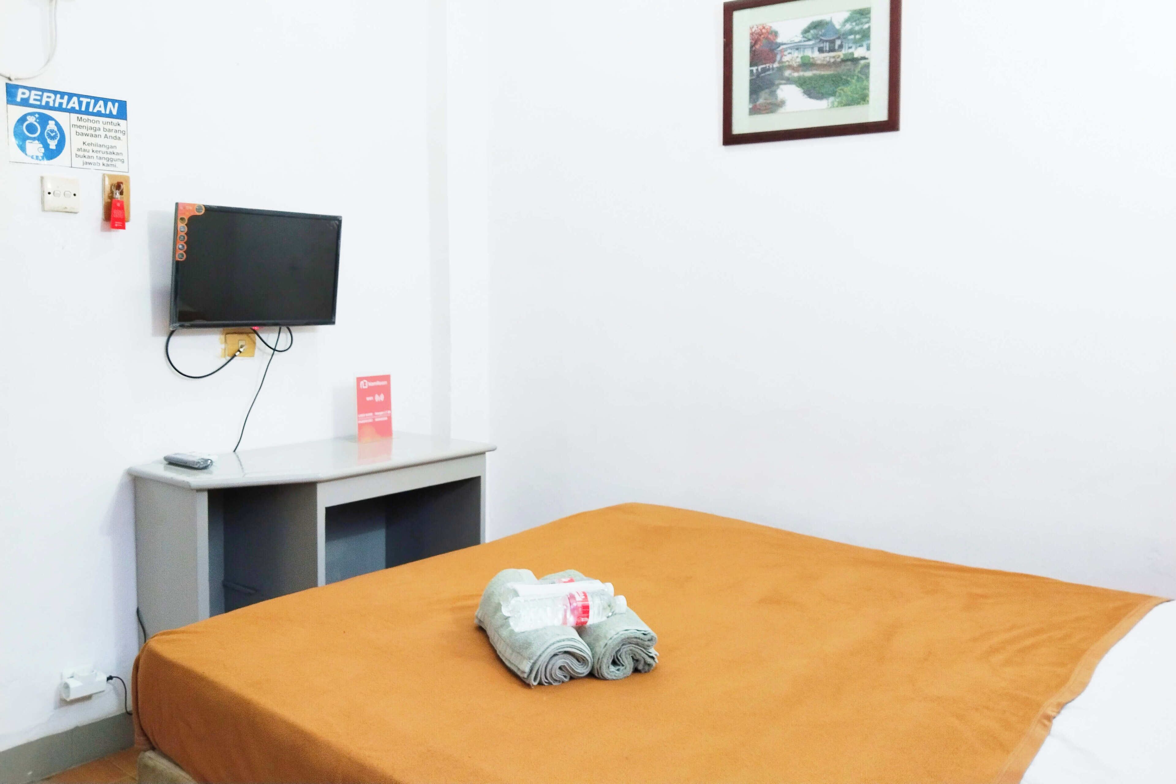 Une chambre d’hôtel avec un lit, un téléviseur fixé au mur, un petit bureau avec une chaise et une serviette pliée posée sur le lit.