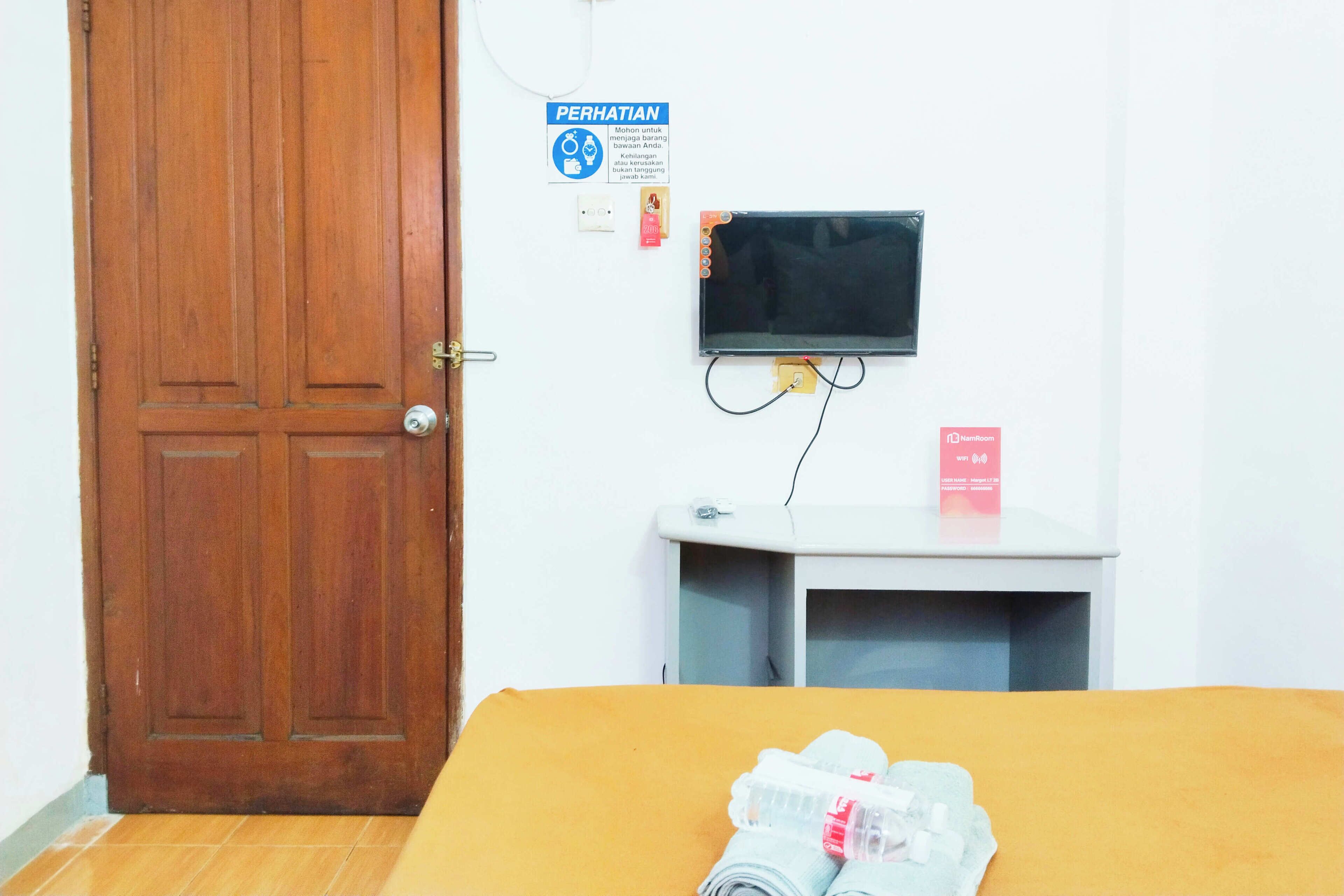 Una habitación con puerta de madera, una cama con colcha amarilla, un televisor sobre un mueble, una mesita blanca con un letrero rojo, y un cartel en la pared que indica servicio de seguridad.