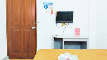 Una habitación con puerta de madera, una cama con colcha amarilla, un televisor sobre un mueble, una mesita blanca con un letrero rojo, y un cartel en la pared que indica servicio de seguridad.