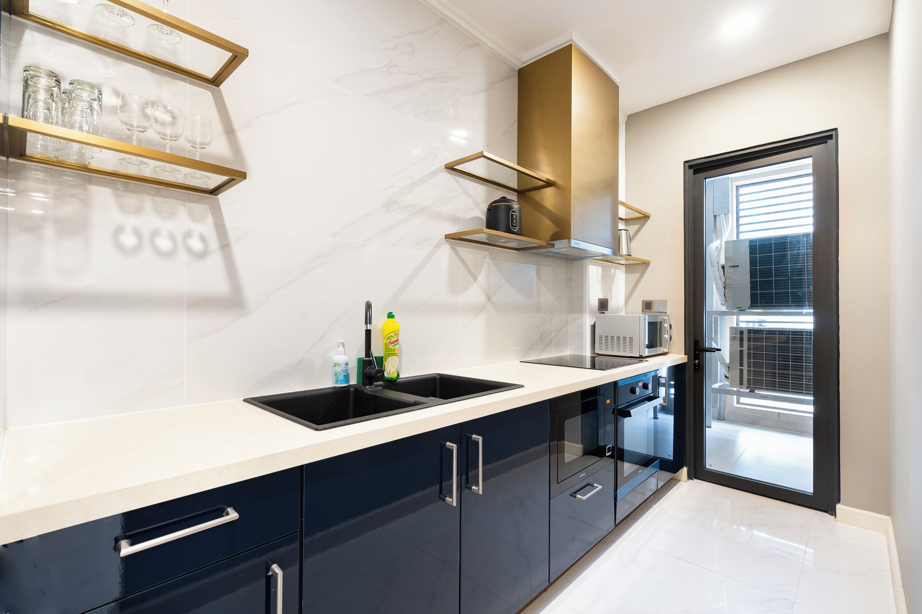 Une cuisine moderne avec un évier noir, un plan de travail blanc et des meubles de rangement sombres. On y trouve un four à micro-ondes, un four traditionnel et une fenêtre avec des stores.