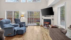 A spacious living room with a high ceiling, large windows, a flat-screen TV, and comfortable seating. - Wooded Cabin Escape w Fire Pit & Family Ready (Berkeley Springs)
