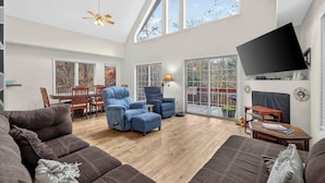 A spacious living room with a high ceiling, large windows, a flat-screen TV mounted on the wall, and a dining area with wooden furniture. - Wooded Cabin Escape w Fire Pit & Family Ready (Berkeley Springs)