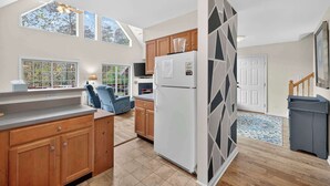 A modern kitchen with wooden cabinets, a white refrigerator, and a dining area with a blue chair. - Wooded Cabin Escape w Fire Pit & Family Ready (Berkeley Springs)