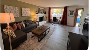 Una sala de estar con sofá, mesa de centro y chimenea. Hay dos cuadros en la pared y una ventana con cortinas.