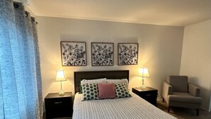 Habitación de hotel con una cama, dos mesitas de noche con lámparas, una silla y tres obras de arte colgadas en la pared.