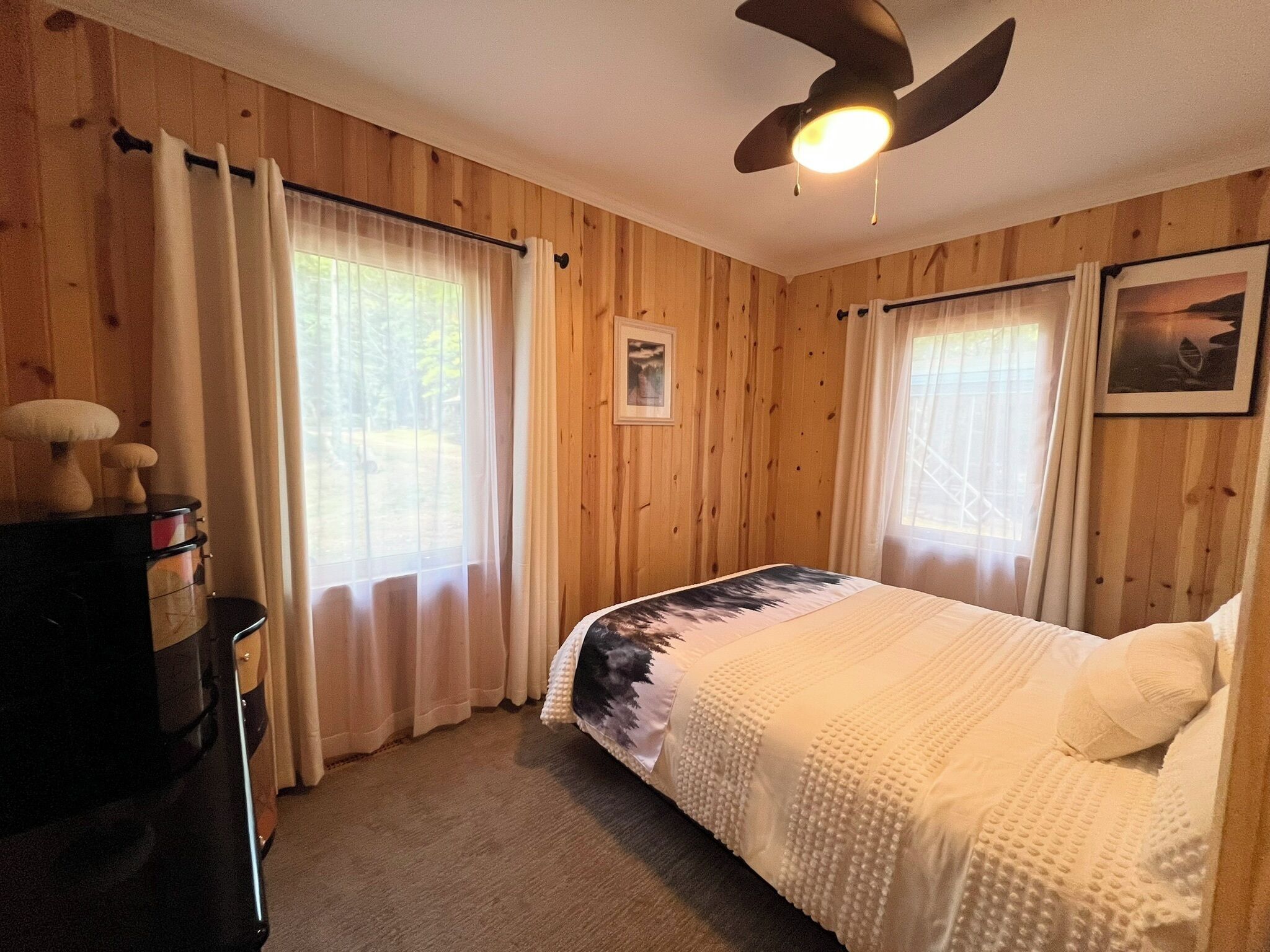 A bedroom with wooden walls, a bed with a white and black patterned bedspread, a ceiling fan, and a dresser with a mirror.