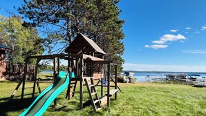 A playground with a slide and a wooden playhouse, set against a backdrop of a lake and boats.