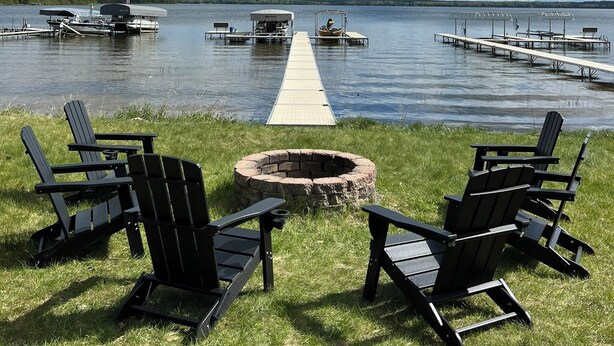 A lakeside view with a dock extending into the water, surrounded by chairs and a fire pit.
