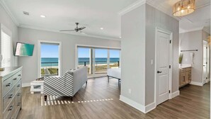 A spacious room with a large window overlooking the beach, a ceiling fan, and a modern light fixture. - The Drift Away (Navarre Beach)