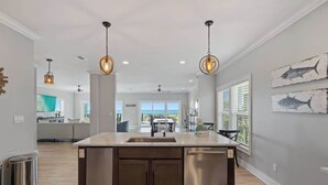 A modern kitchen with a central island, stainless steel appliances, pendant lighting, and a view to the dining area. - The Drift Away (Navarre Beach)