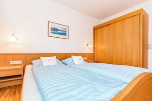 A hotel room with a wooden headboard, two beds with blue bedding, a nightstand, a wall-mounted lamp, and a framed picture on the wall.