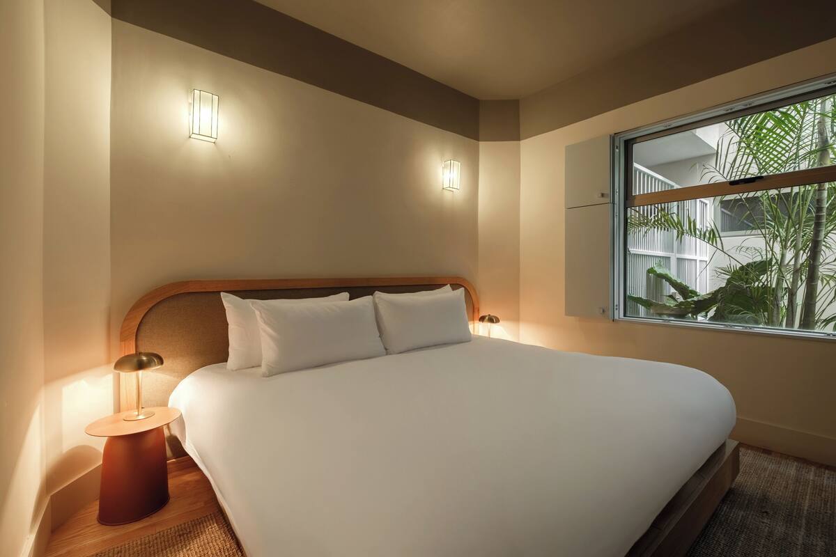 A neatly made bed with white linens, two wall-mounted lights, and a window with a view of greenery.