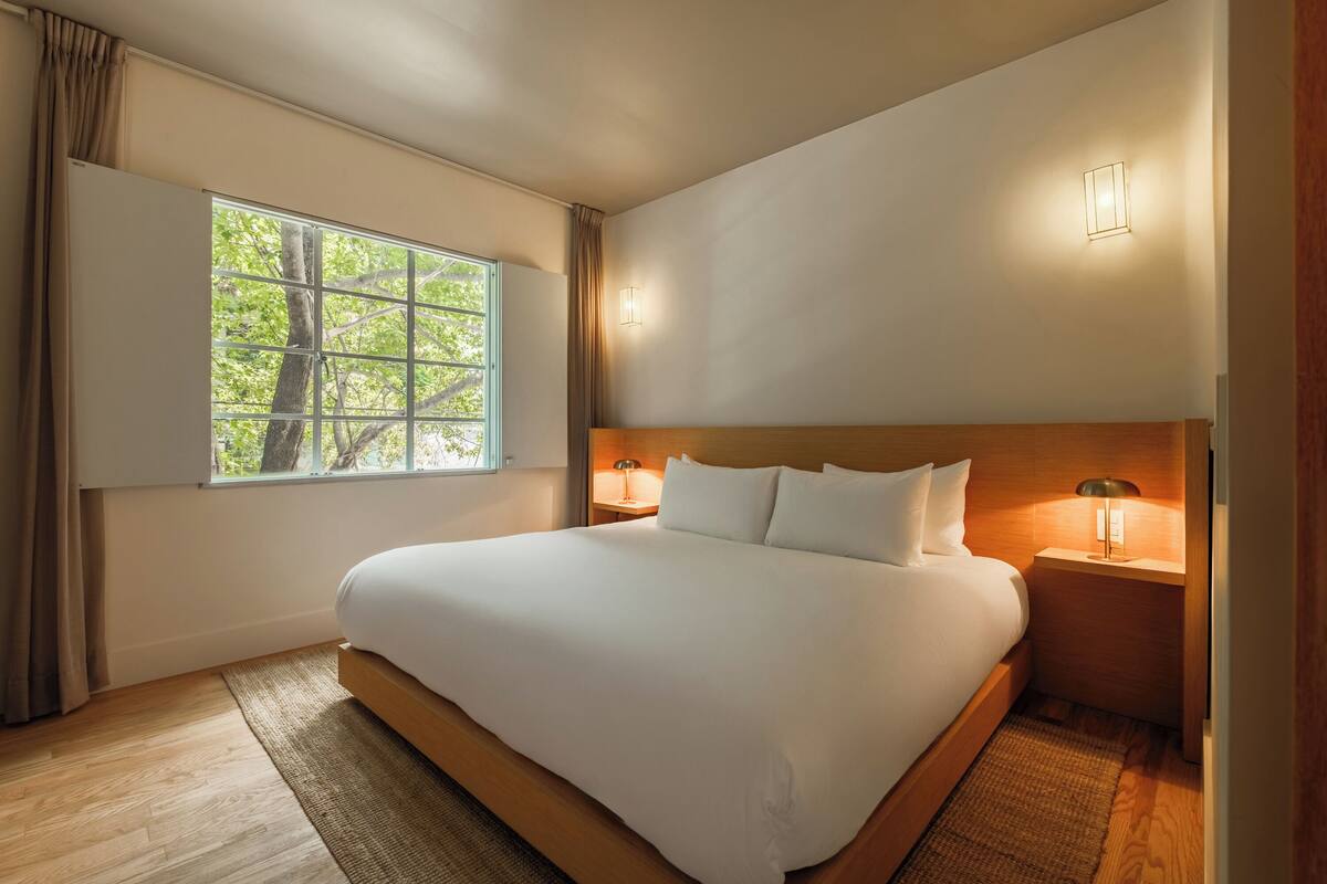 A large bed with a wooden headboard, two bedside tables with lamps, a window with curtains, and a view of trees.