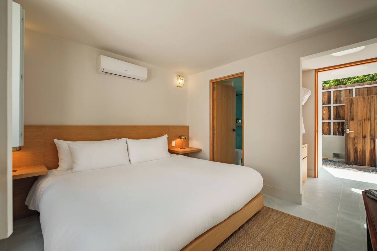 A bedroom with a large bed, wooden headboard, bedside tables, an air conditioning unit, and a view of a wooden fence through an open door.