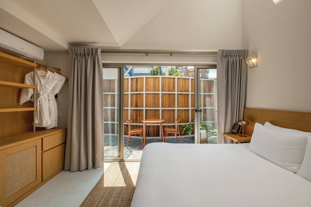 A hotel room with a bed, a wooden wardrobe, a table with chairs, and a view through a glass door.