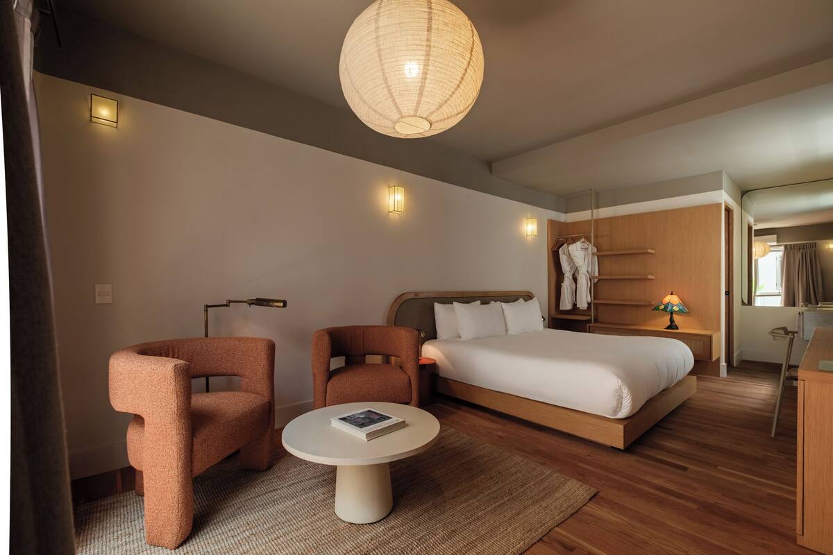 A modern hotel room with a bed, two armchairs, a round table, and a hanging light fixture.