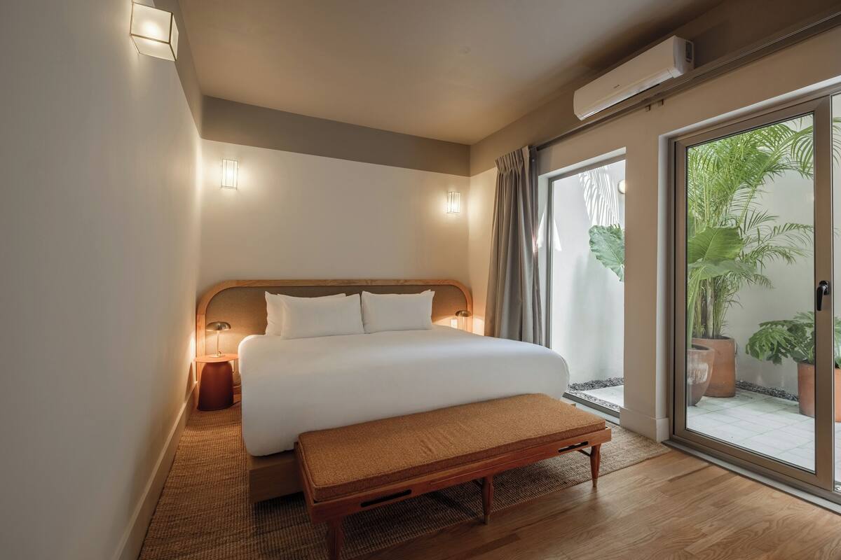 A hotel room with a large bed, bedside lamps, a bench, and a sliding glass door leading to a balcony with potted plants.