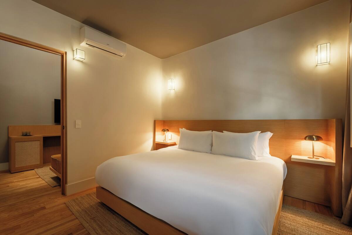 A hotel room with a large bed, two bedside tables with lamps, a wooden headboard, a mirror, and an air conditioning unit.