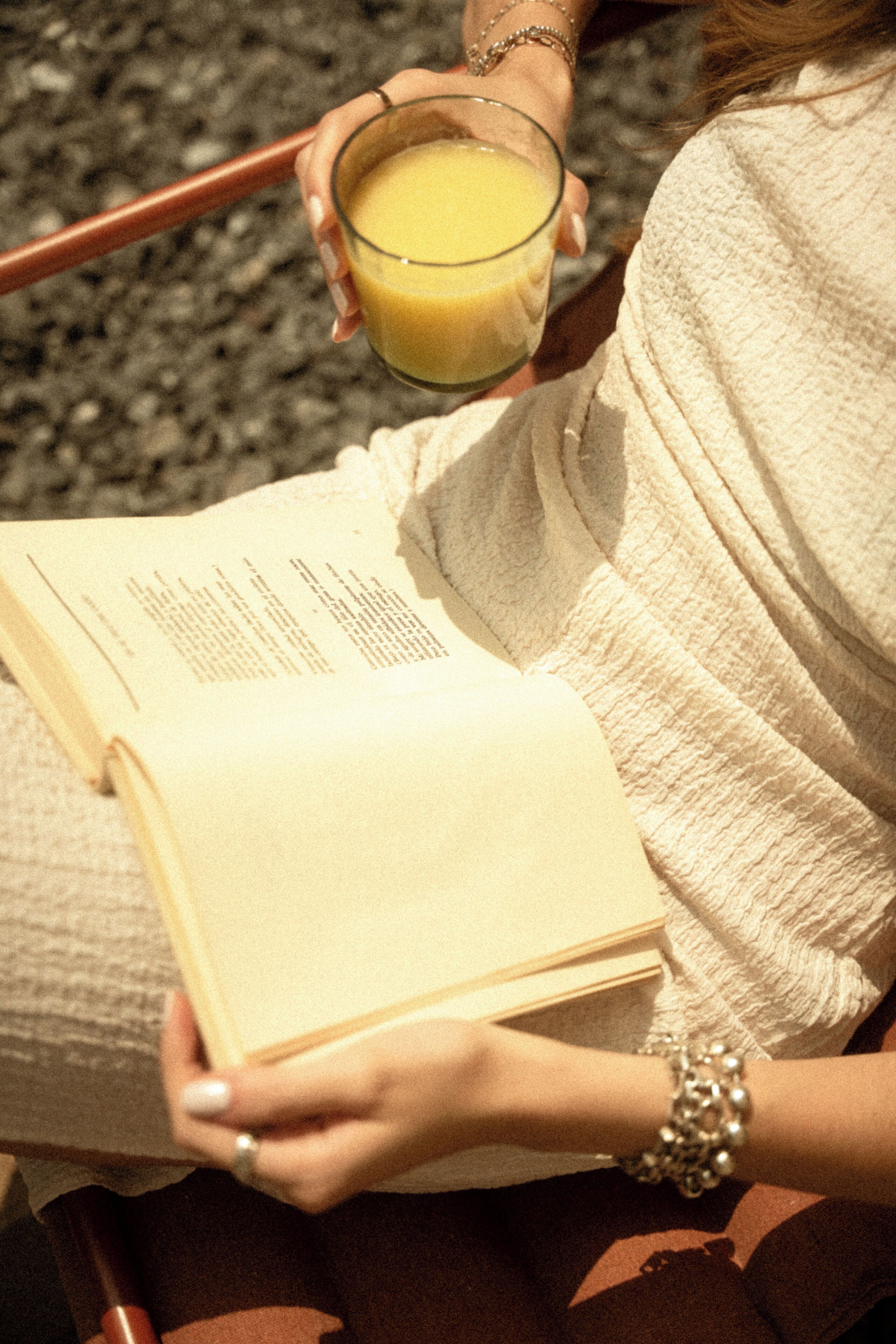 A person reading a book while holding a glass of orange juice.