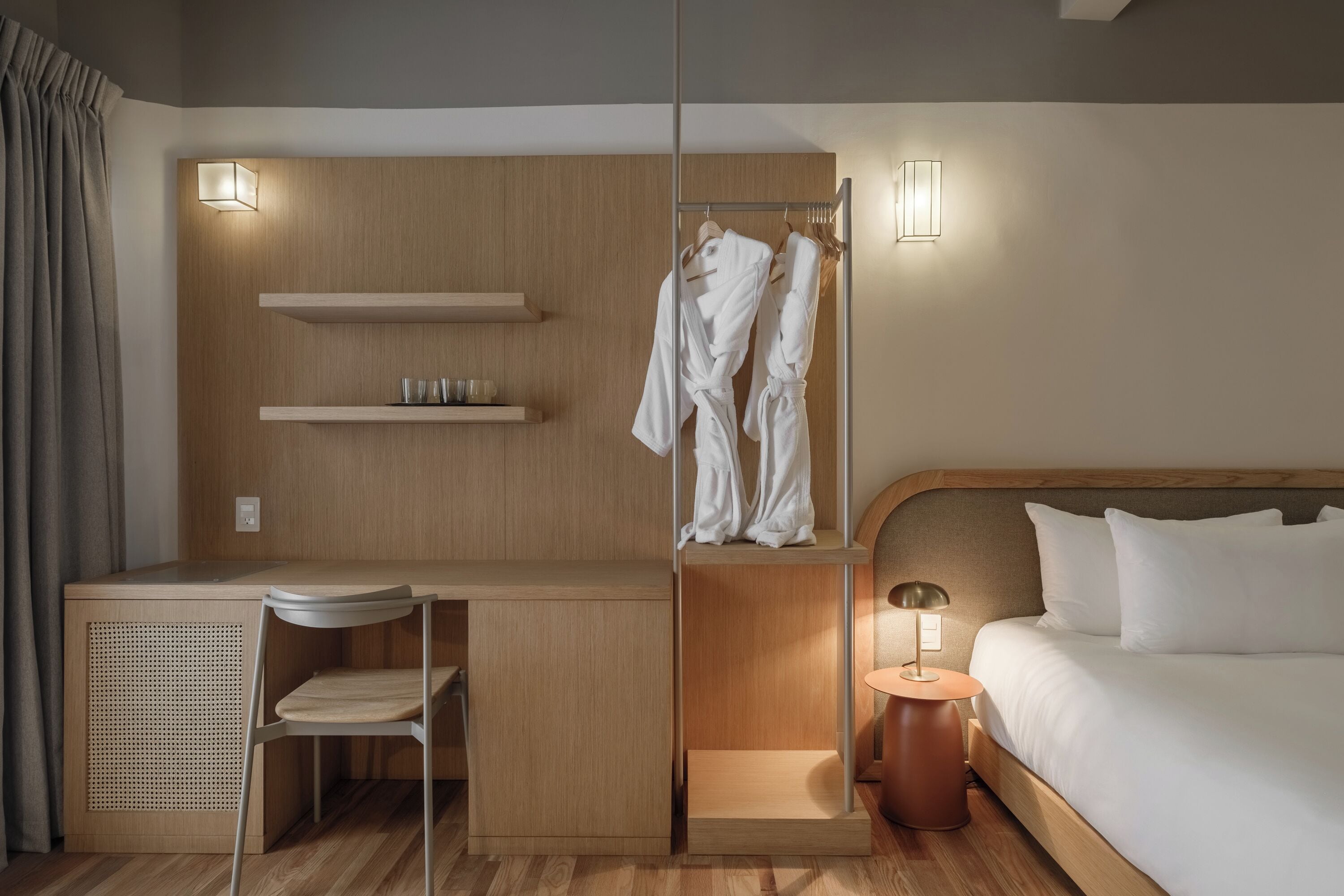 A hotel room with a wooden desk, a chair, a bed with white bedding, a bedside table with a lamp, a wardrobe with hanging robes, and two wall-mounted light fixtures.