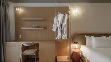 A hotel room with a wooden desk, a chair, a bed with white bedding, a bedside table with a lamp, a wardrobe with hanging robes, and two wall-mounted light fixtures.