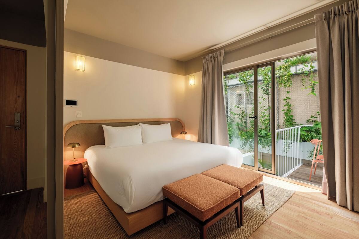 A hotel room with a large bed, a small table, a bench, and a view of a balcony with plants.