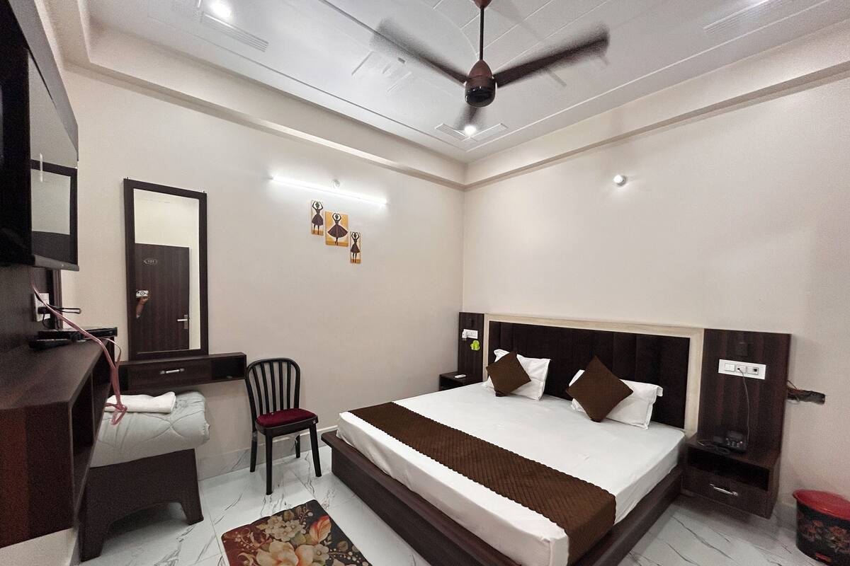 Une chambre d’hôtel comprenant un lit, une table de chevet, une chaise, un bureau, un miroir, un ventilateur de plafond et une petite poubelle.