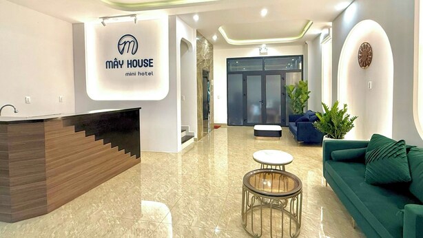 A modern hotel lobby with a reception desk, a seating area with a green sofa, and a large glass door entrance. - Mây House Grandworld on the beach (Phu Quoc)