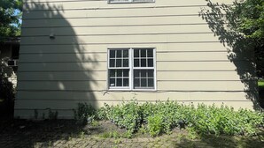 A two-story building with a symmetrical facade, featuring a central arched window and a smaller rectangular window below. The building has a flat roof and is surrounded by a paved area with small plants.
