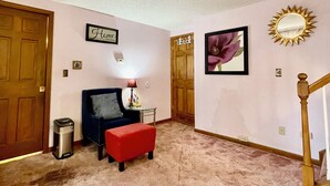 A hallway with a door labeled "Home," a framed flower picture, a lamp, a trash can, and a red ottoman.