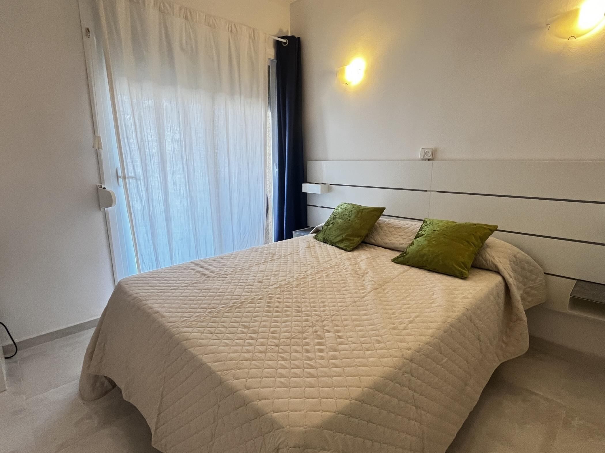 A bedroom with a bed, a headboard, a window with curtains, and a wall-mounted light.
