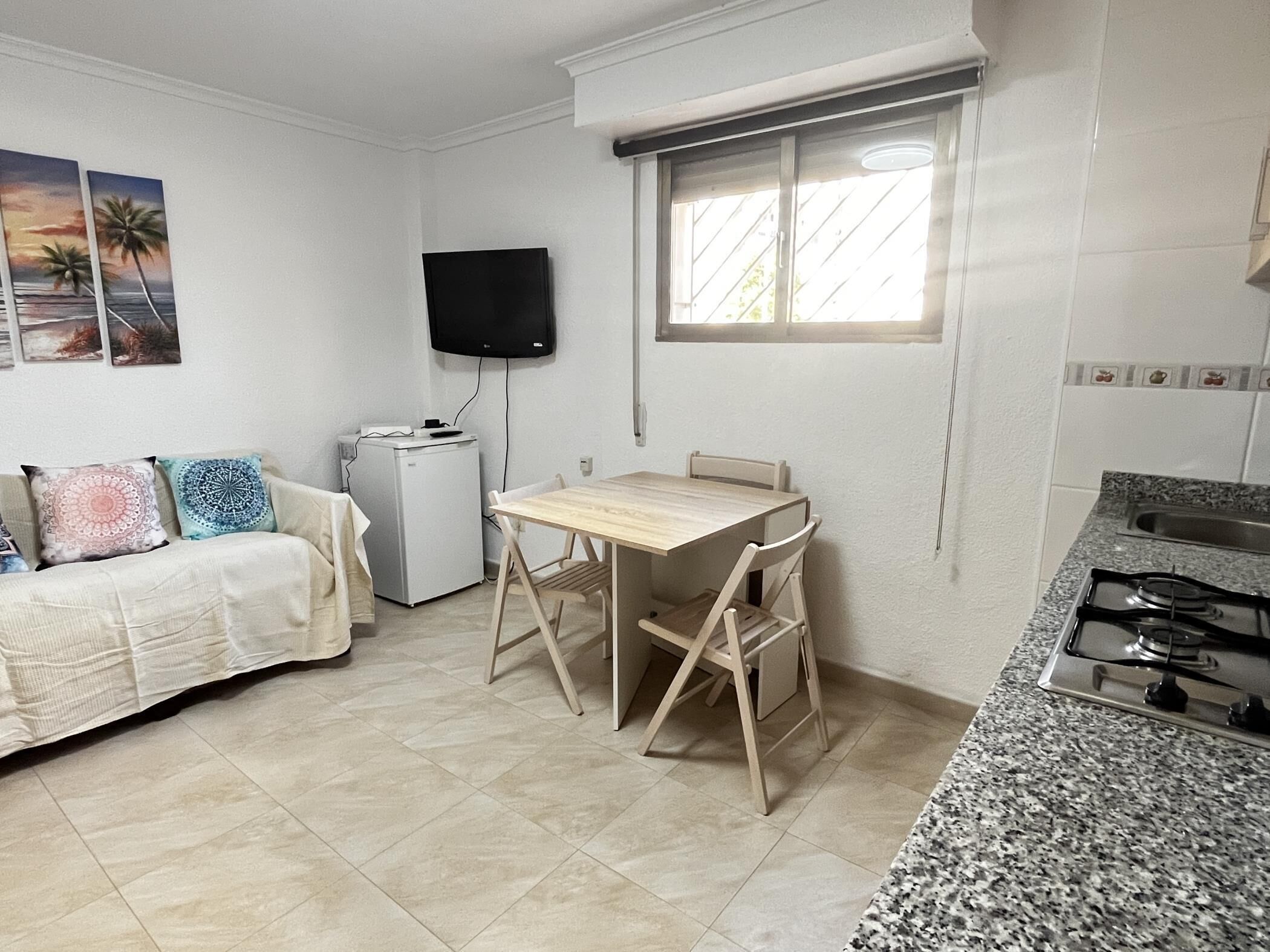 Een knus woongedeelte met een kitchenette, een eettafel, een bank en een aan de muur bevestigde televisie.